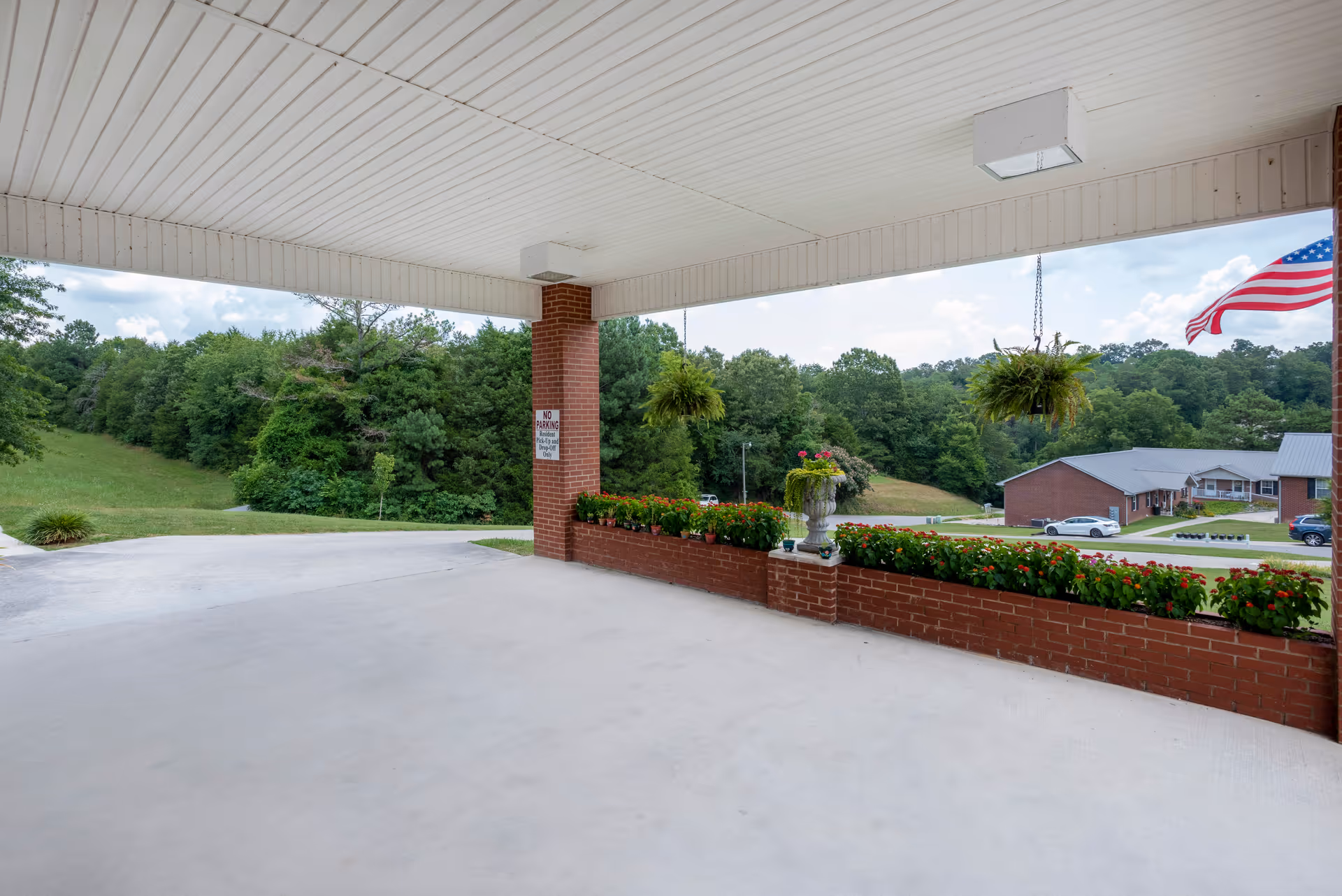 Covered entrance/porte-cochere with brick planters and hanging ferns overlooking a lawn, trees, and a neighboring building.