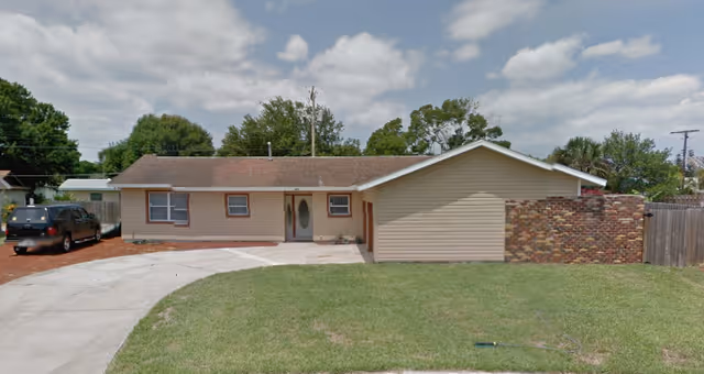 Single-story beige house with driveway, lawn, and brick wall in front yard.