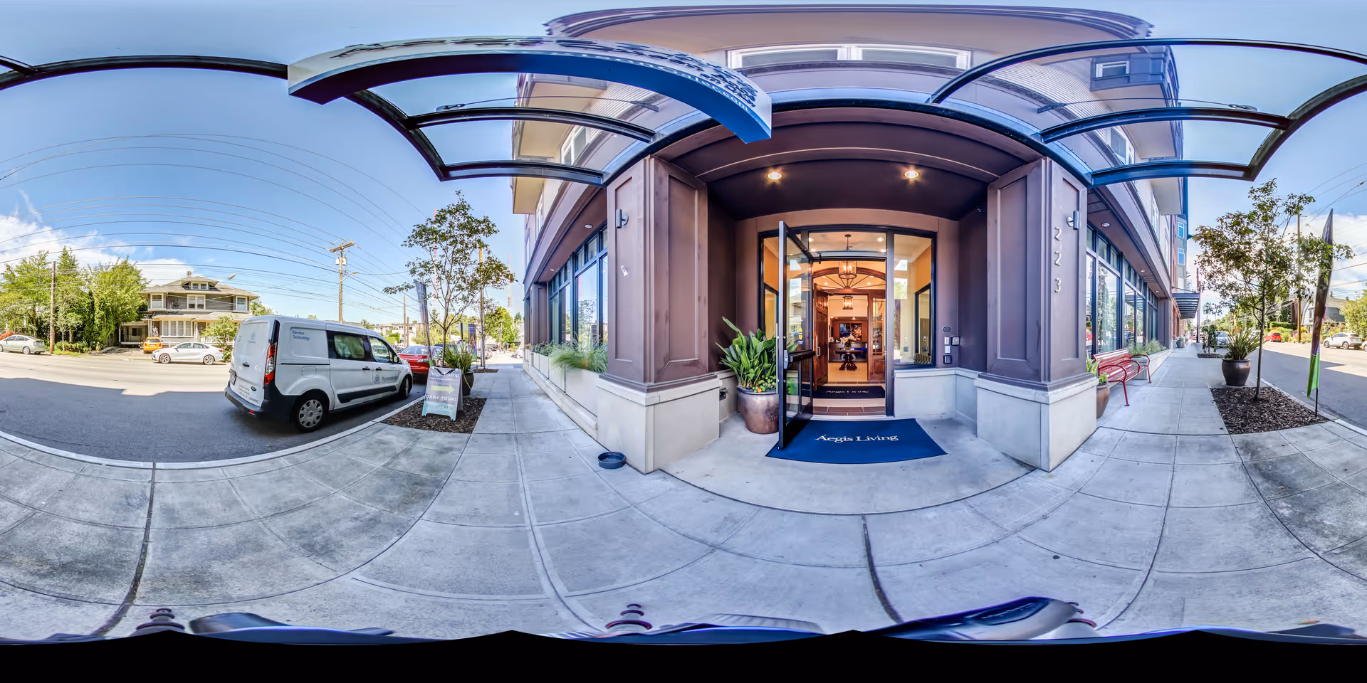 Exterior entrance of Aegis Living Queen Anne Galer facility with a glass door open, a blue welcome mat with the facility name, potted plants on either side of the entrance, and a covered walkway. The sidewalk and street with parked vehicles and trees are visible under a clear sky.