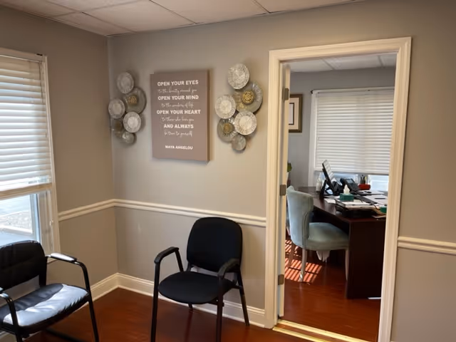 A small waiting area with two black chairs along a gray hallway and office doorway.
