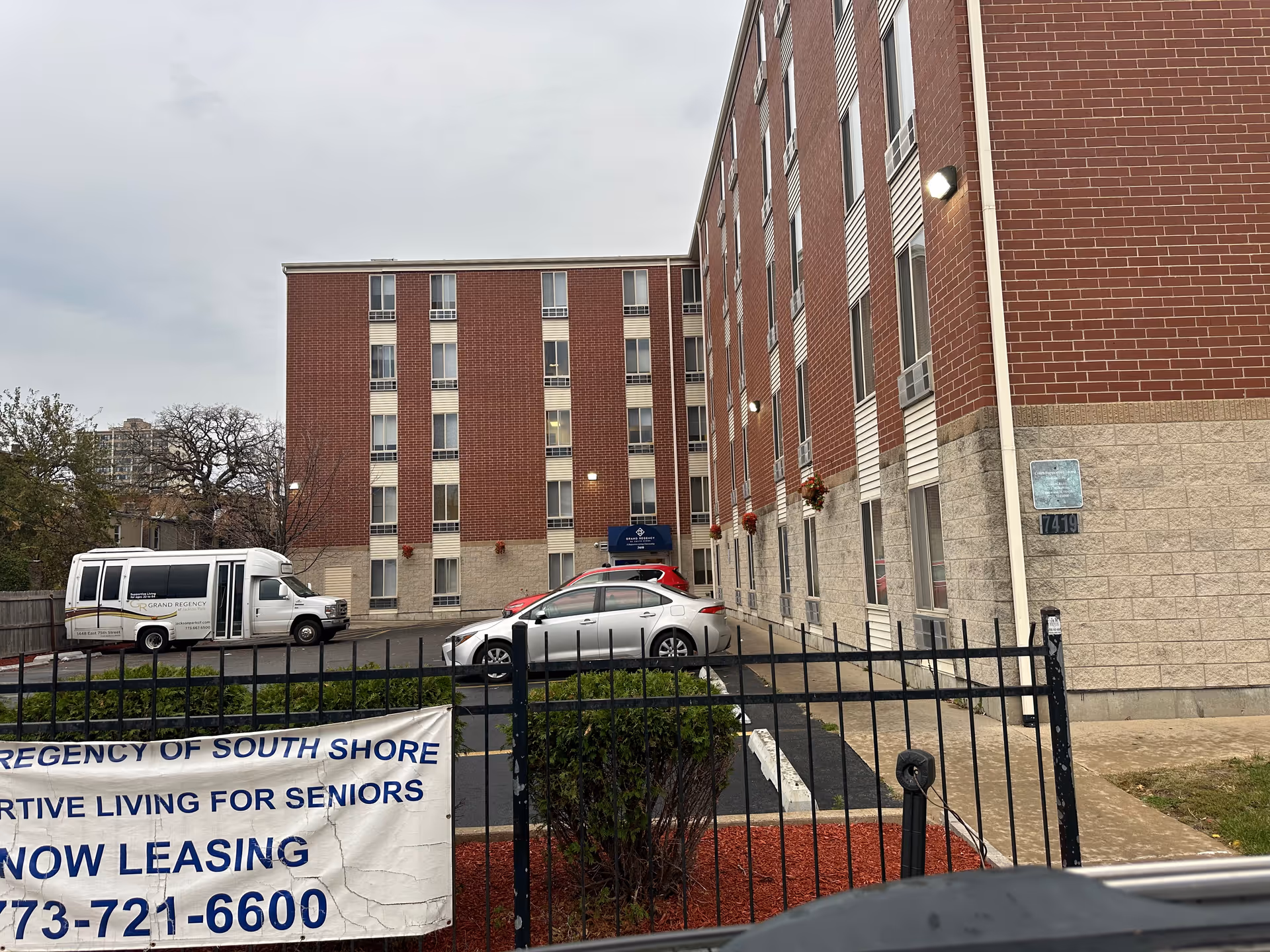 Exterior view of a multi-story brick building with several windows and a parking lot in front. There are a few parked cars and a white shuttle van with the logo 'Grand Regency' on the side. A black metal fence with a banner advertising supportive living for seniors and leasing information is visible in the foreground. The sky is overcast.