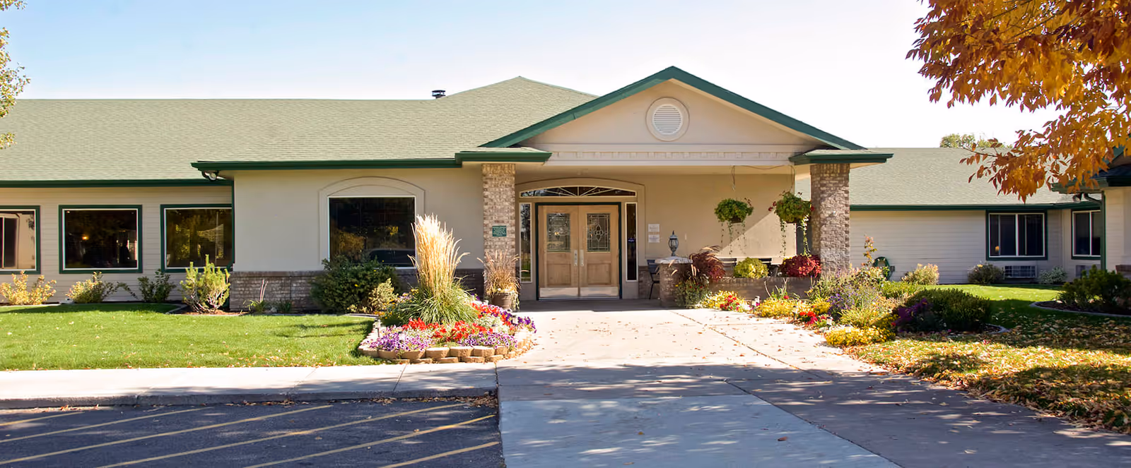 Front exterior view of a single-story building with a green roof and beige walls, featuring a central entrance with double glass doors, surrounded by landscaped flower beds and green grass under a clear sky.