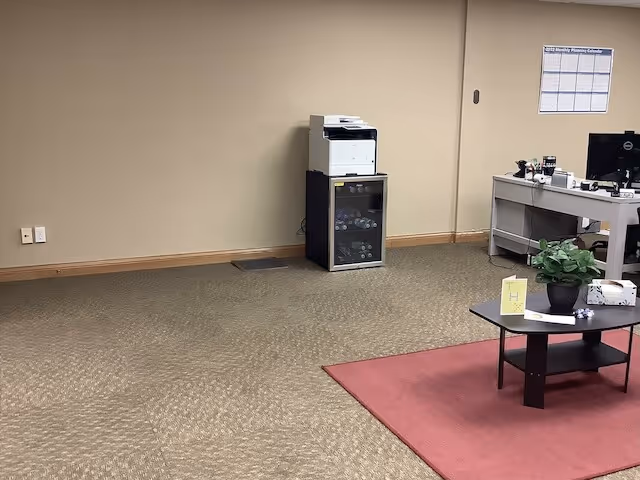 Reception area inside a facility with desk, computer, printer, plant, and red rug.