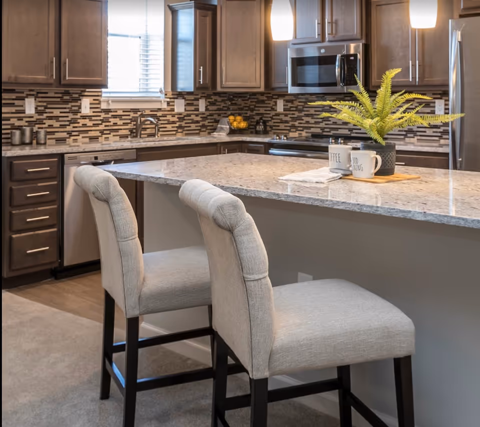 Bright modern kitchen featuring a large island with a granite countertop, two upholstered bar stools, pendant lights, and dark wood cabinets.
