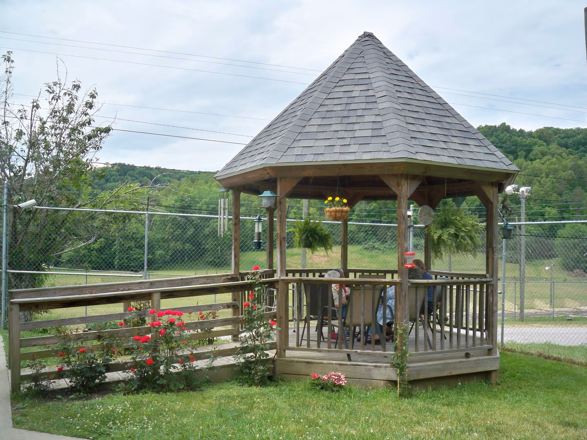 A wooden gazebo with a shingled roof situated outdoors on a grassy area. Inside the gazebo, several people are seated around a table. The gazebo is surrounded by a wooden railing and has hanging plants and flowers nearby. In the background, there is a chain-link fence and a view of green trees and hills under a cloudy sky.