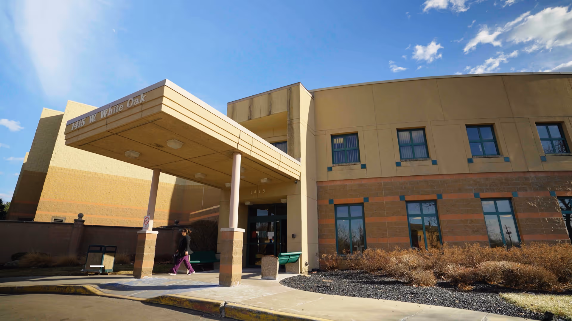 Exterior view of a two-story building with a covered entrance supported by two columns. The building has beige and brown walls with several windows. Two people are walking near the entrance under a clear blue sky with some clouds.