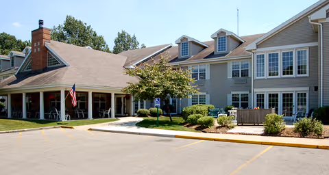 Exterior view of a senior living facility building with beige siding, multiple windows, and a covered entrance with an American flag. There is a small landscaped area with bushes and a tree near the entrance, and a parking lot in the foreground.