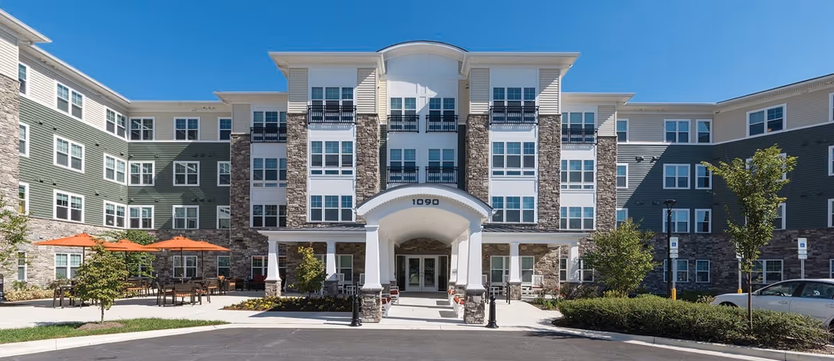 Front exterior of a multi-story senior apartment building with a covered entrance, outdoor seating with orange umbrellas, and landscaped driveway.