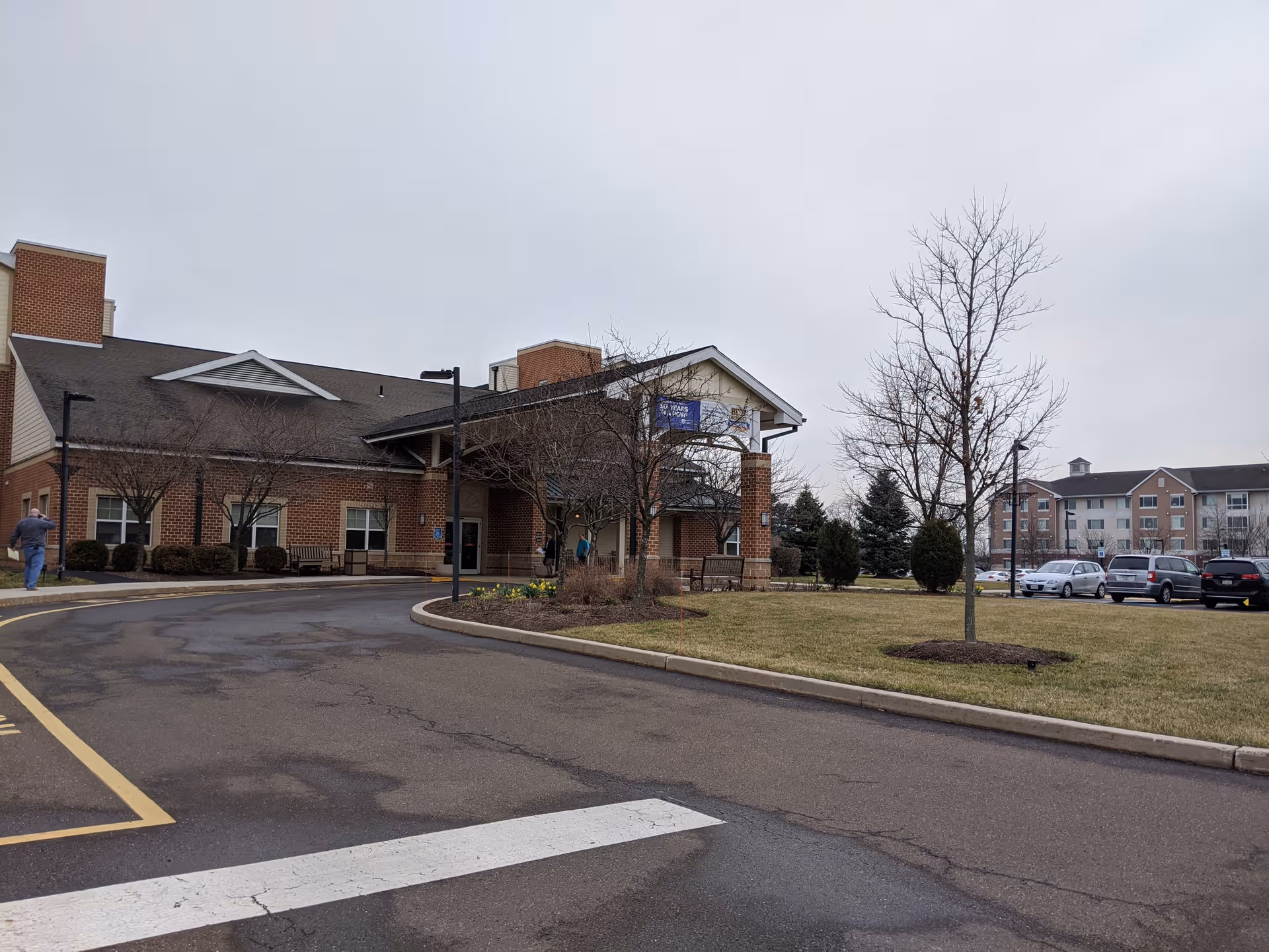 Brick senior living building with entrance canopy, parking lot, and leafless trees under gray sky.