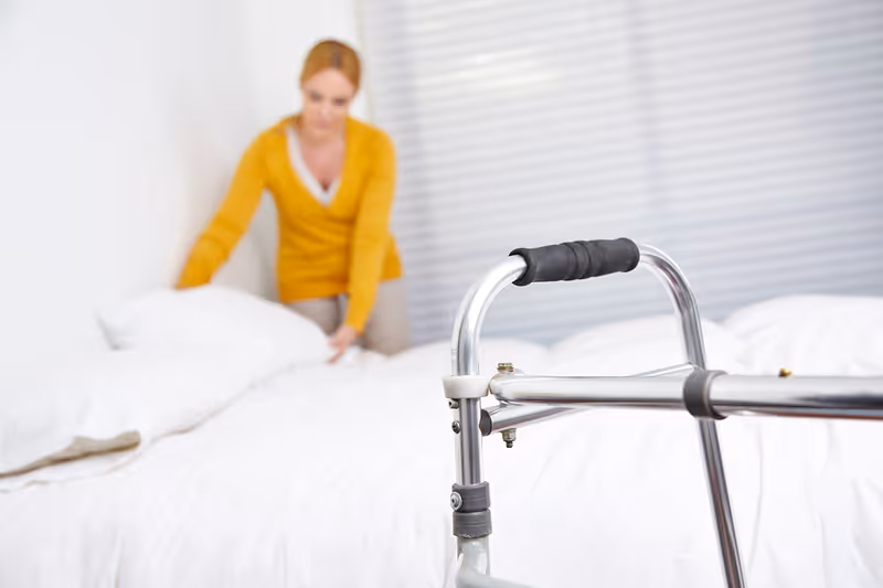 A caregiver arranges pillows on a bed in a bright, homey bedroom with a walker nearby.