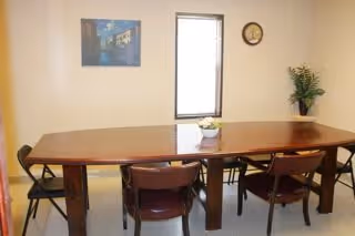 A small dining area with a rectangular wooden table surrounded by six chairs. On the table is a small decorative centerpiece. The room has a window with blinds, a wall clock, a framed picture, and a potted plant on a side surface.