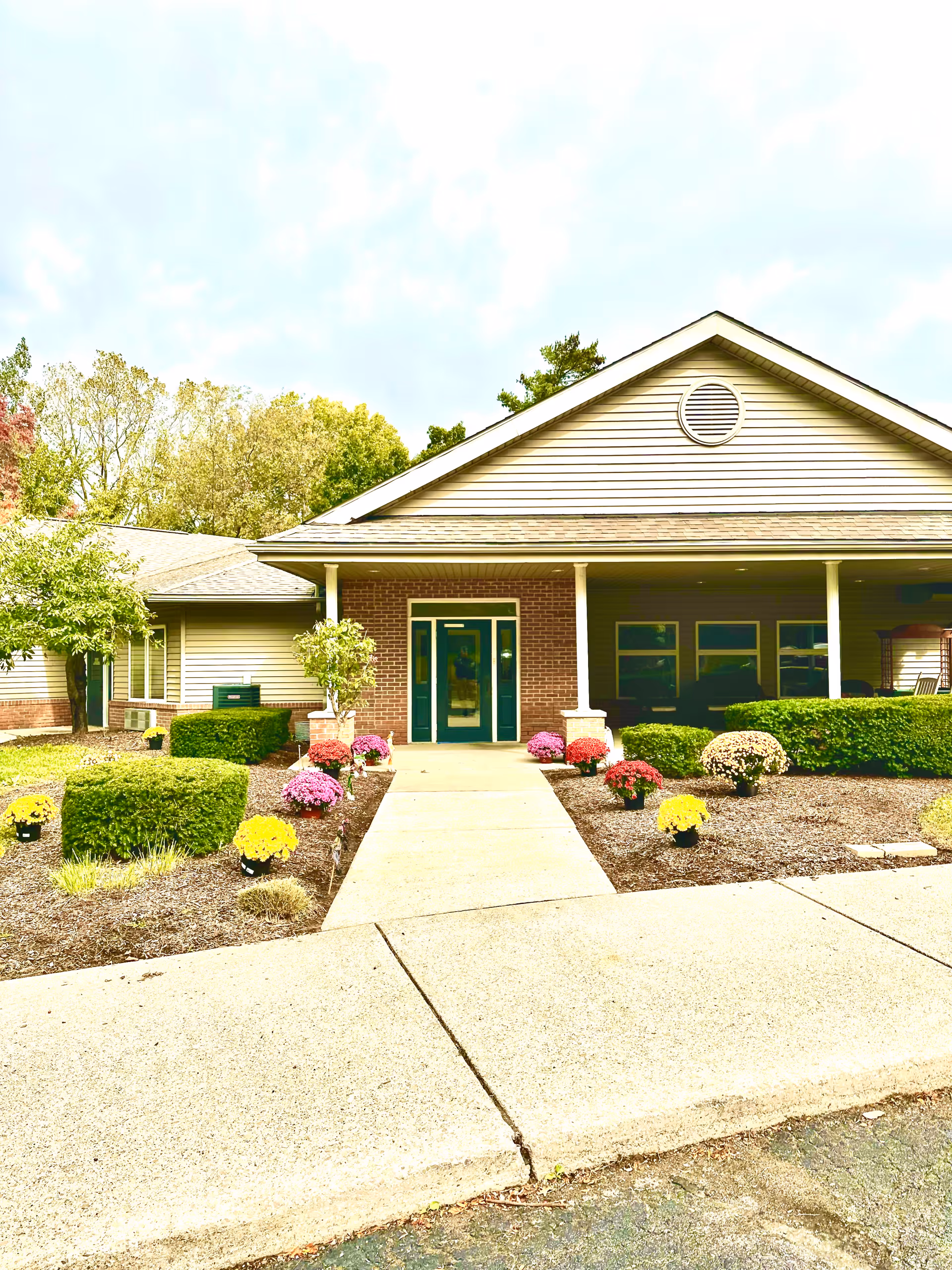 Front exterior view of a single-story building with a gabled roof, a covered entrance supported by columns, and a walkway leading to double doors. The landscaping includes neatly trimmed bushes and colorful flowers along the walkway.