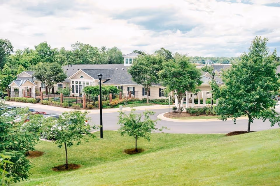 Exterior view of a senior living facility building surrounded by green trees and a well-maintained lawn, with a circular driveway and parked cars visible.