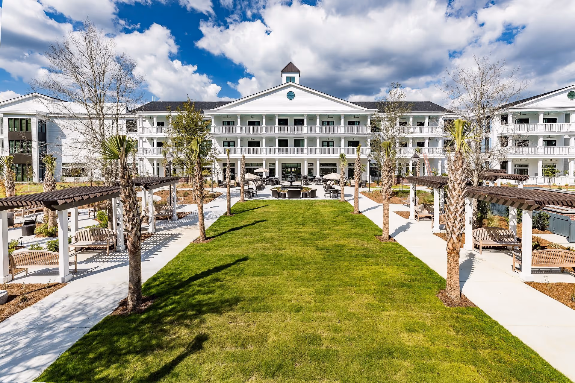 Outdoor view of a senior living facility with a large green lawn in the center, flanked by paved walkways and pergolas with benches. The building in the background is white with multiple balconies and a central cupola under a partly cloudy blue sky.