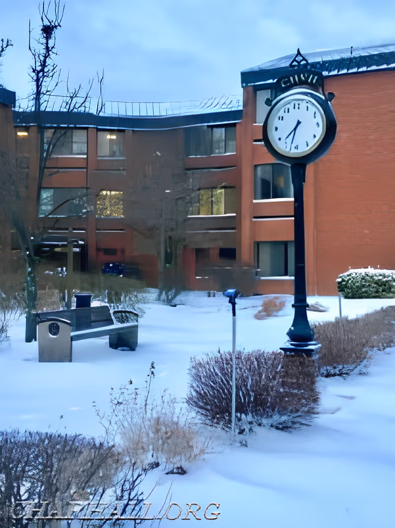 Snow-covered courtyard with a tall street clock, bench, shrubs, and a brick building in the background.