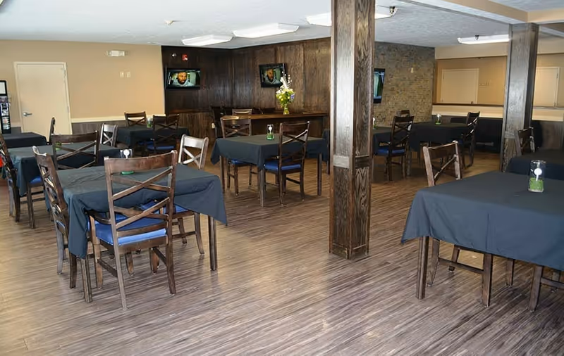 A dining area with multiple wooden tables and chairs covered with dark tablecloths. The room has wooden flooring, beige walls, and two wooden pillars. There are two small wall-mounted televisions on a dark wood-paneled wall, and a vase with flowers is placed on a counter in the background.