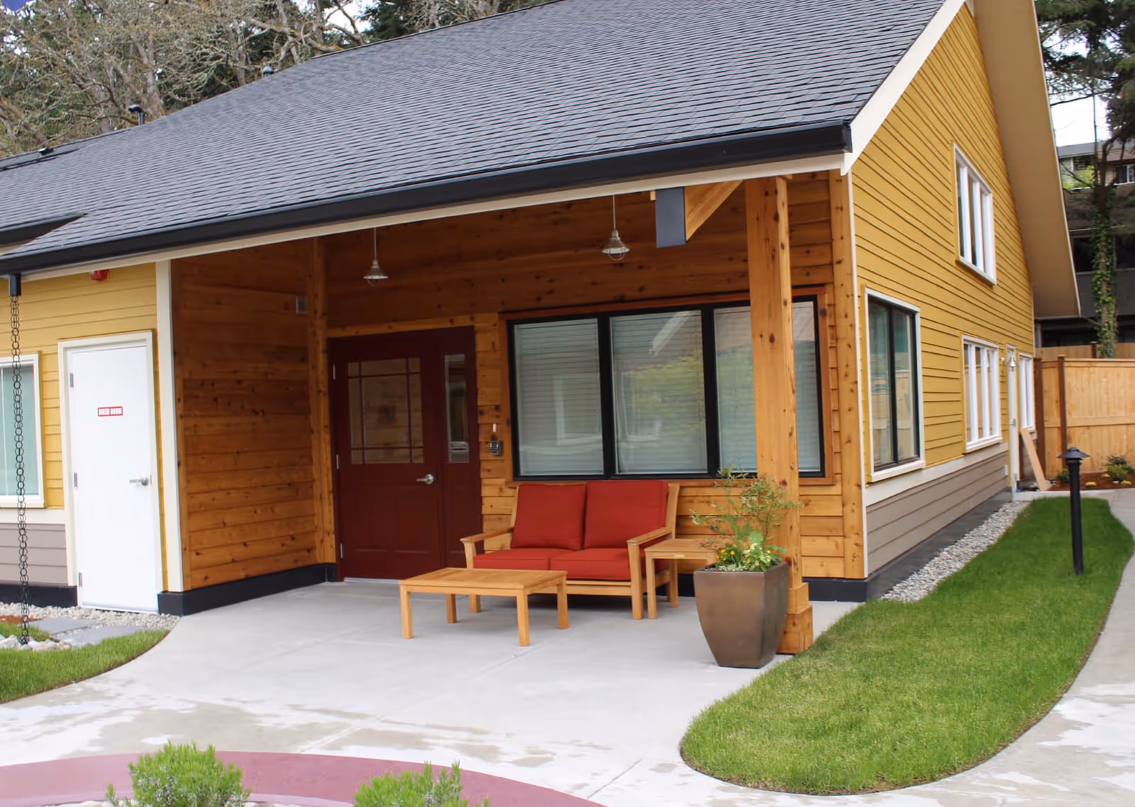 Covered entrance of a yellow and wood-clad cottage with a red door, red cushioned outdoor seating, potted plant, and a small lawn.