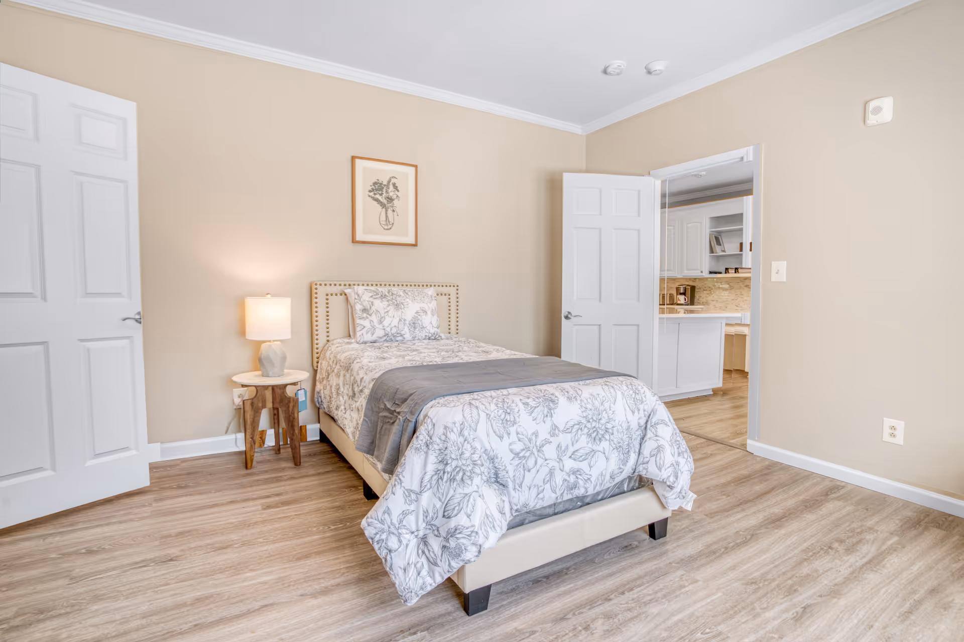 Cozy bedroom with a neatly made bed, nightstand, lamp, and doorway to an adjacent kitchen.