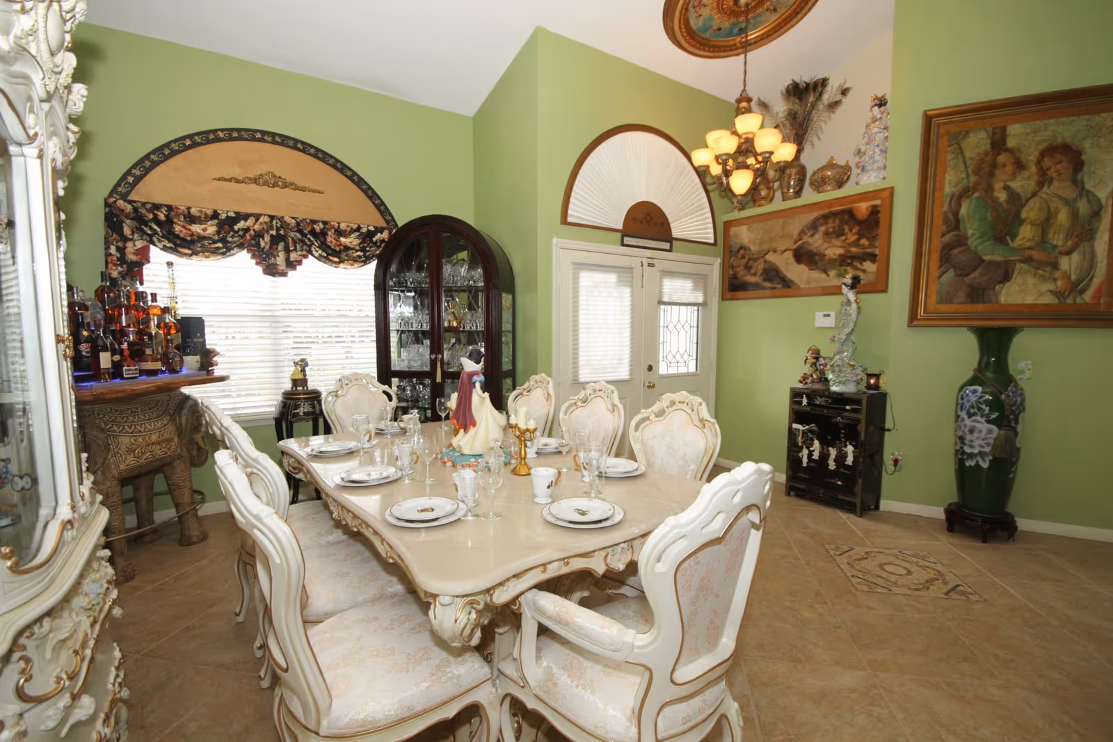 Elegant dining room with ornate table, matching chairs, chandelier, and china cabinet against green walls.