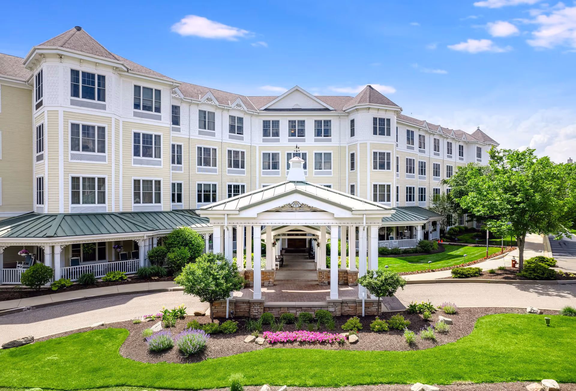 Exterior view of a large, multi-story senior living facility with white and light yellow siding, numerous windows, and a covered entrance with white pillars. The building is surrounded by well-maintained landscaping including green grass, bushes, and flower beds under a blue sky with some clouds.