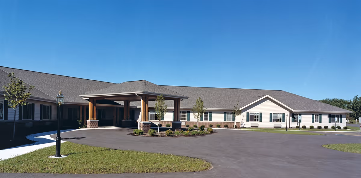 Exterior view of Ridgestone Village, a single-story building with a covered entrance supported by wooden pillars, surrounded by a paved driveway, small landscaped areas with young trees, and a clear blue sky.