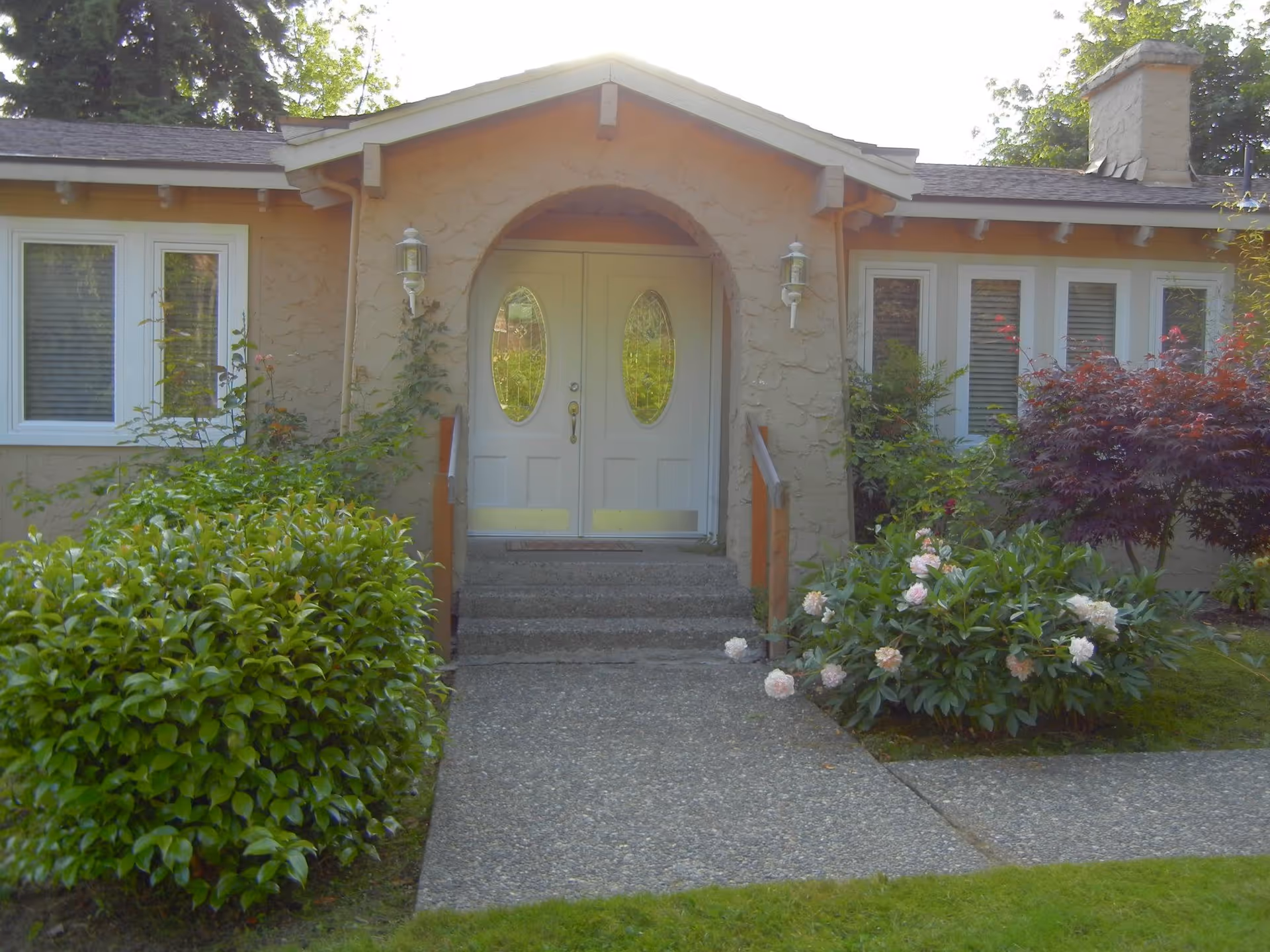 Front entrance of a beige building with double doors, steps, and landscaped shrubs.