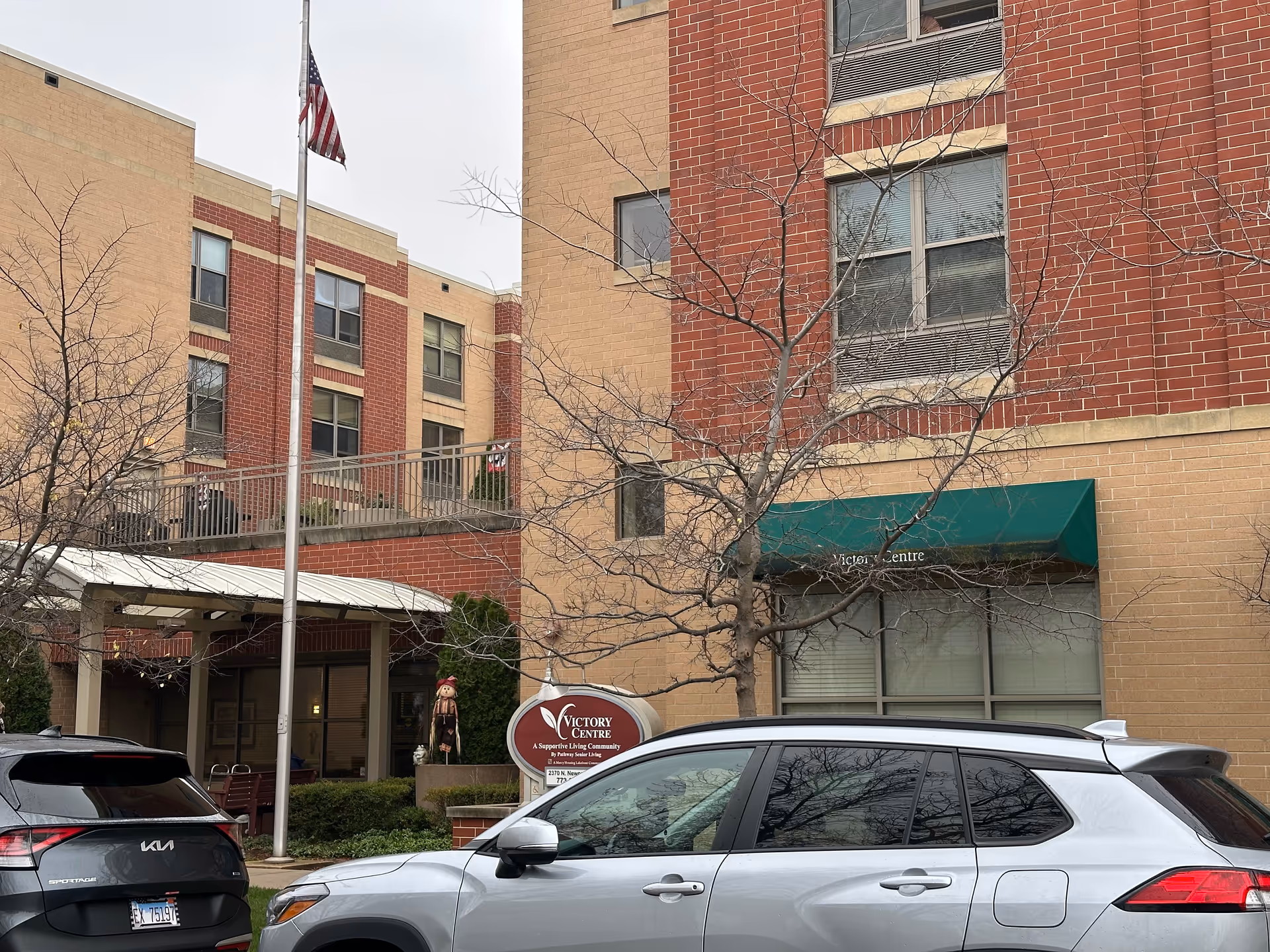 Exterior view of Victory Centre of Galewood, a multi-story brick building with a green awning over a window. There is a flagpole with an American flag, a sign for the facility, and two parked cars in front. Leafless trees are visible near the entrance.