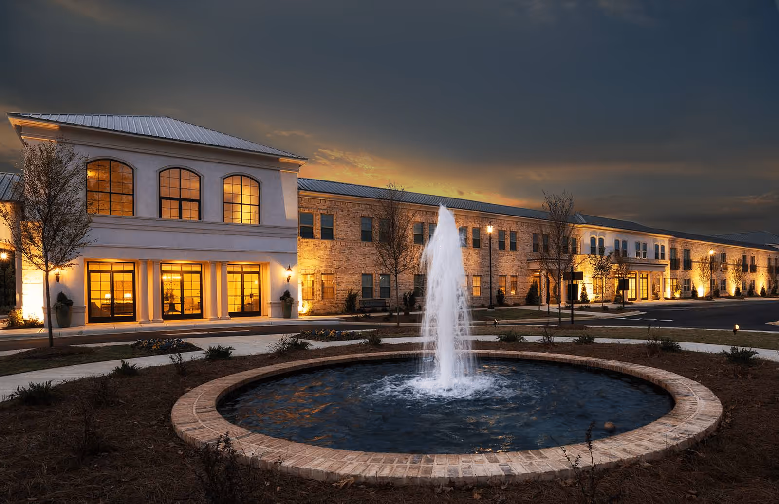 Evening exterior view of The Blake at Flowood with illuminated windows and a circular fountain.