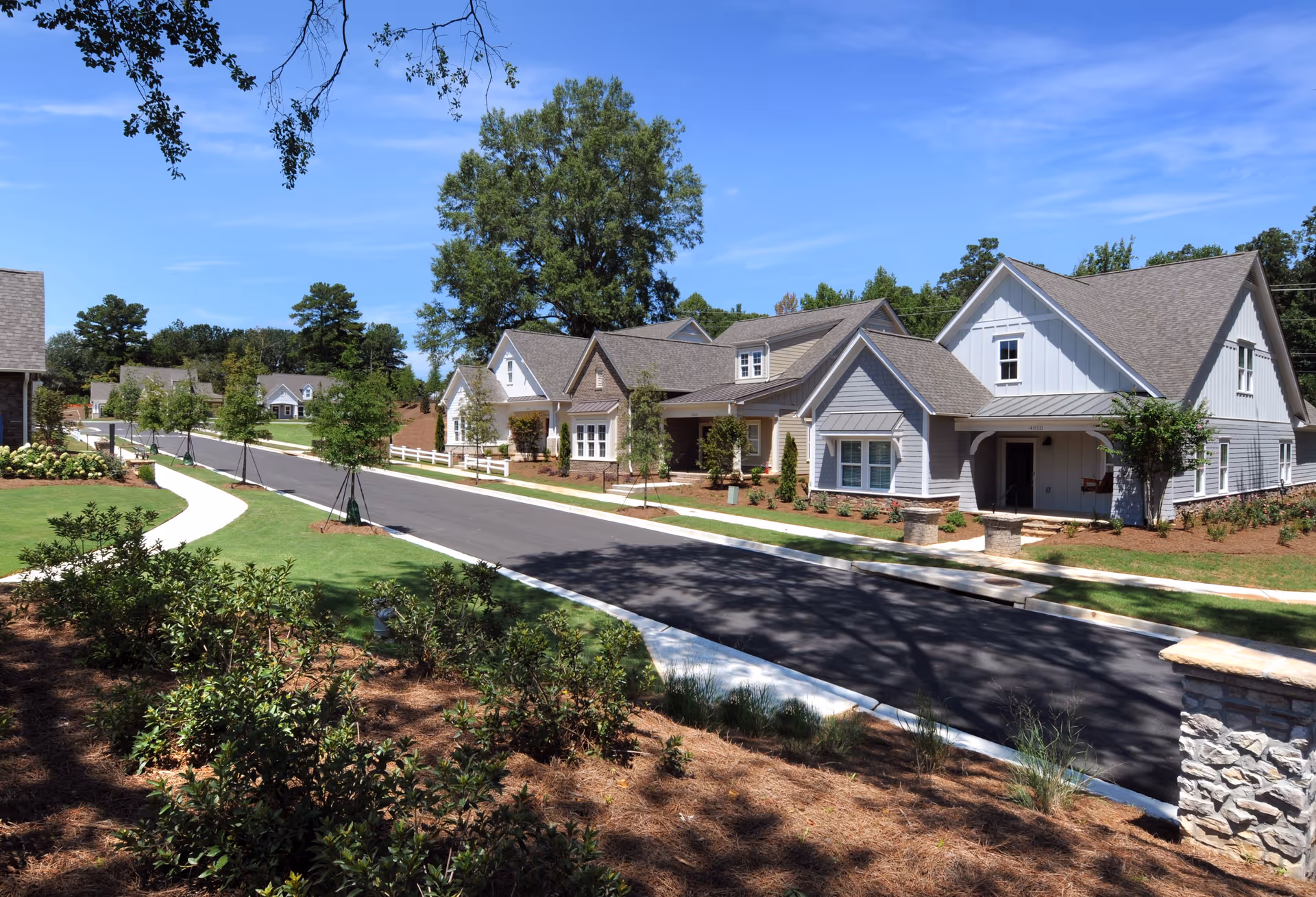 A sunny street view of a residential community with several single-story houses featuring pitched roofs, manicured lawns, young trees, and landscaped gardens under a clear blue sky.