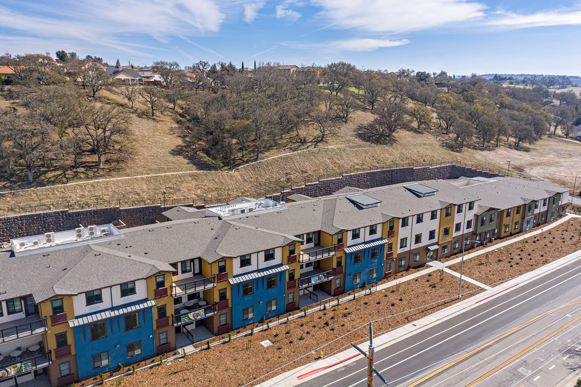 Aerial view of a three-story apartment-style building with colorful facades along a road in front of a grassy hillside.