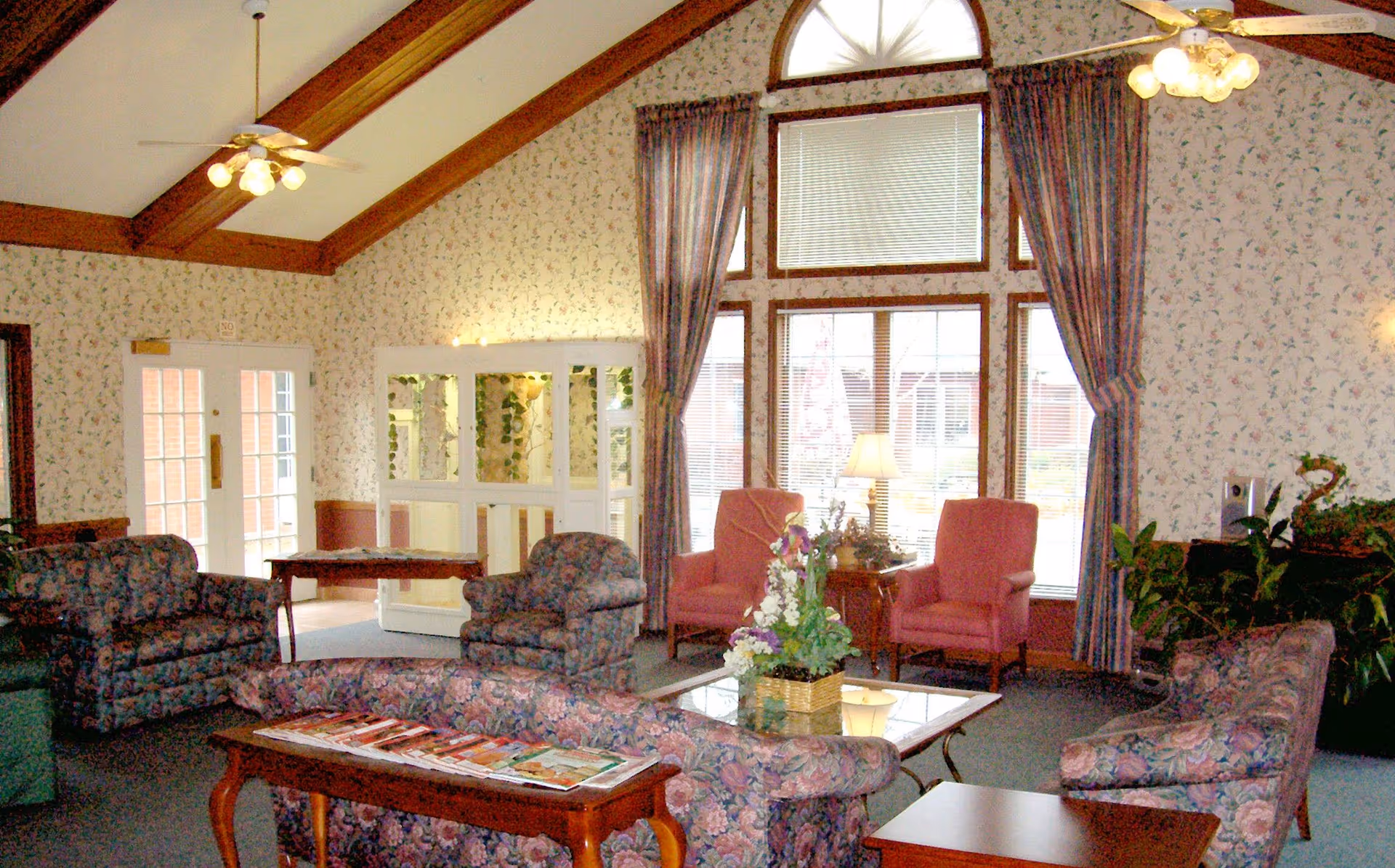 A cozy living room area in Seminary Village with floral patterned sofas and armchairs arranged around a glass coffee table with a flower arrangement. The room features high vaulted ceilings with wooden beams, large windows with blinds and floral curtains, and two pink upholstered chairs near a side table with a lamp. The walls have floral wallpaper, and there are ceiling fans with lights.