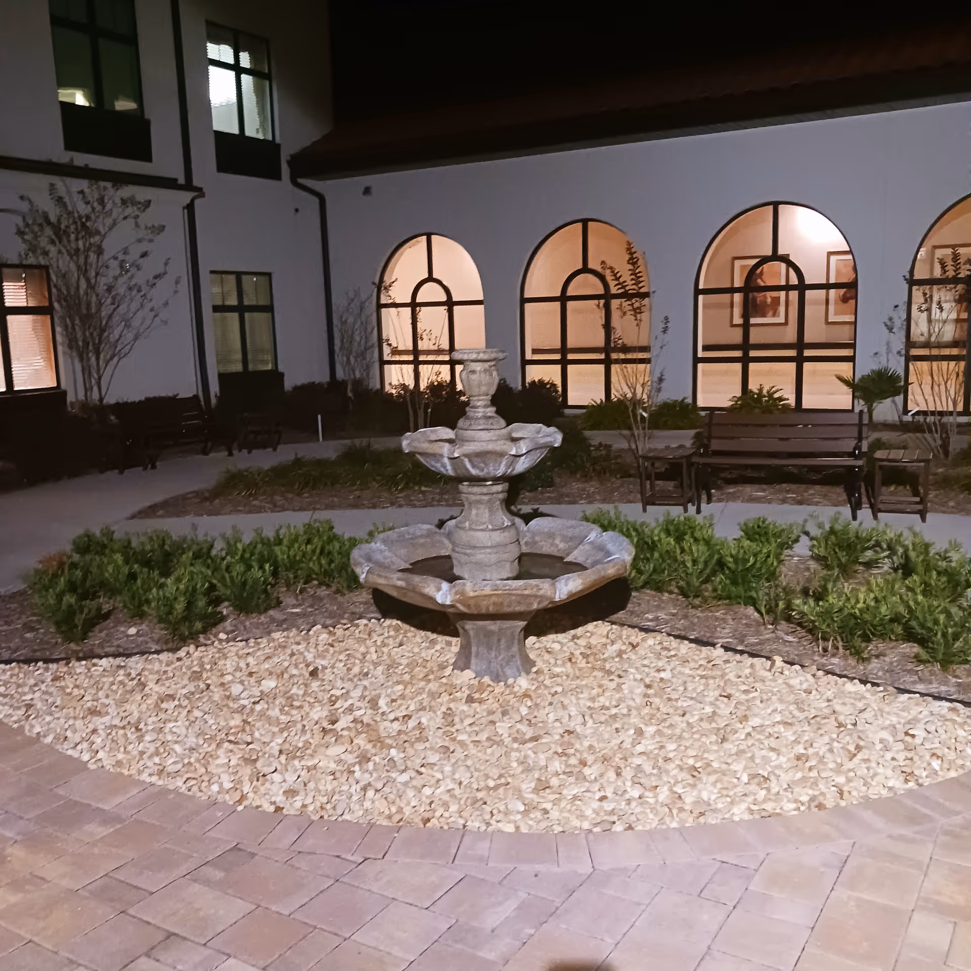 Night courtyard with a stone tiered fountain, benches, and arched windows.