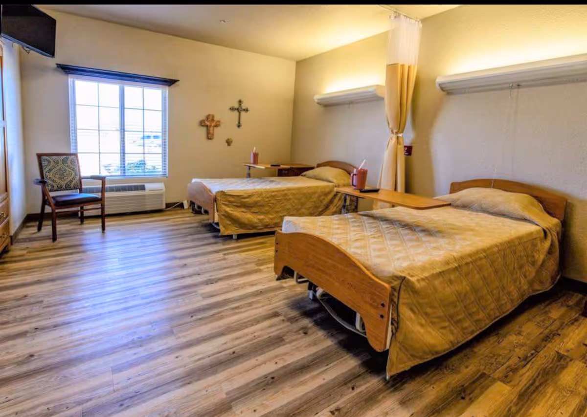 A room with two single beds covered in beige quilts, each with a wooden headboard and a small overbed table. There is a wooden chair with patterned upholstery near a large window with blinds. The walls are light-colored with three crosses hanging on one wall. The floor is wood-patterned laminate, and there is a curtain divider between the beds.