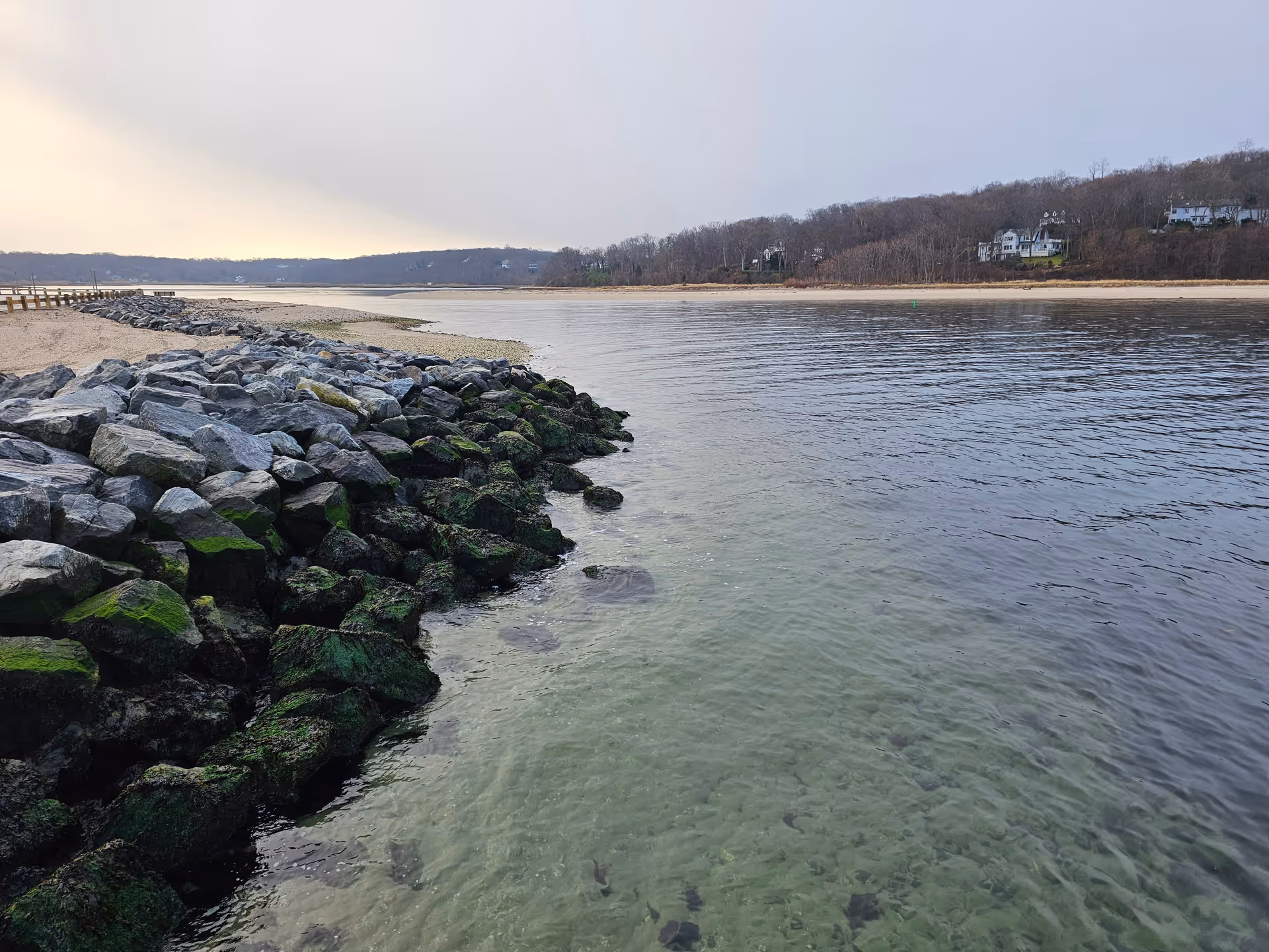 Coastal shoreline with rocky breakwater, clear shallow water, and distant houses on a hillside.