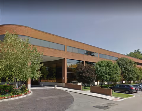 Brick senior living facility exterior with curved entrance, landscaped trees, and parked cars.