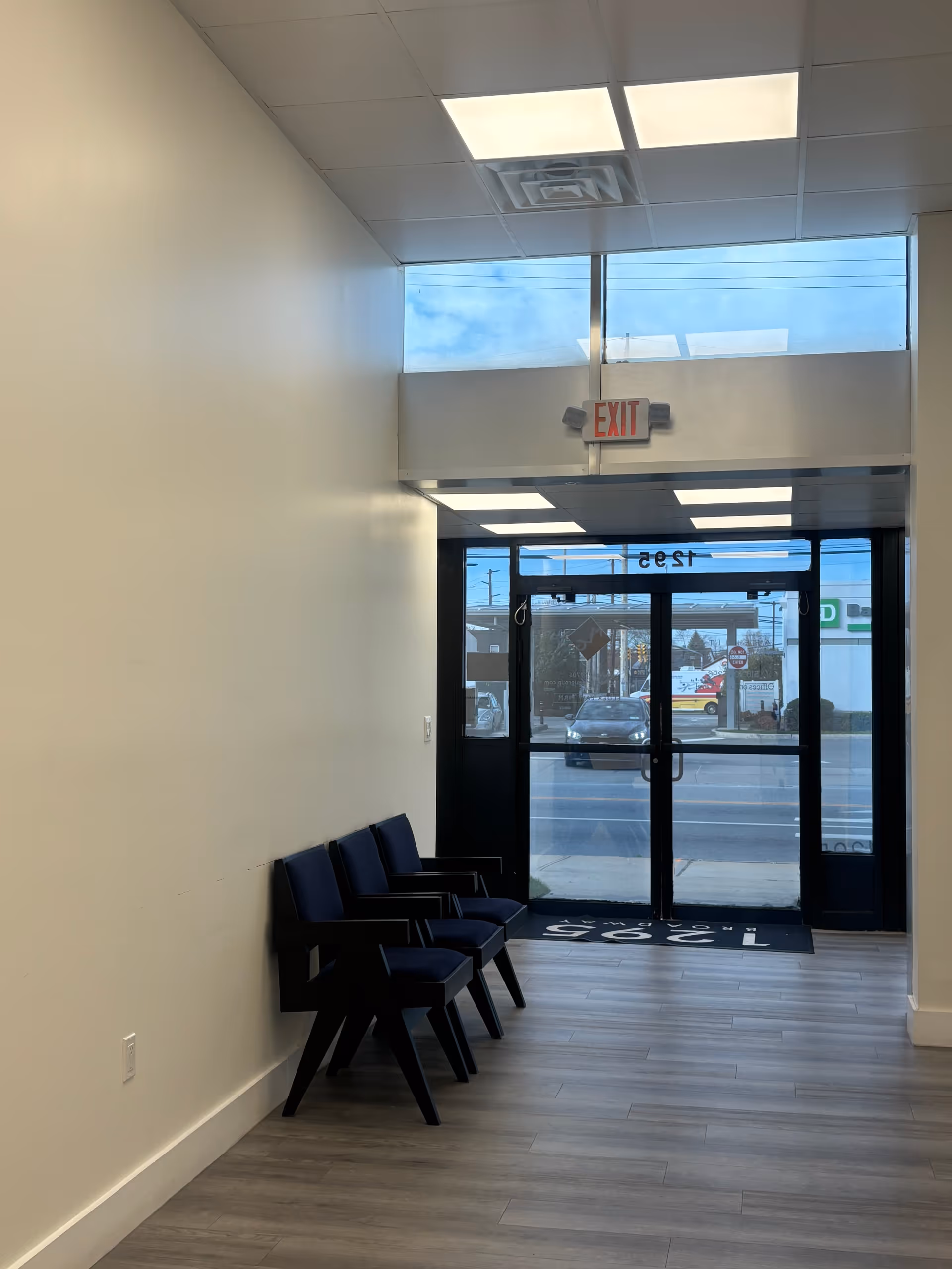 Interior hallway and waiting area with blue chairs near glass entrance doors.