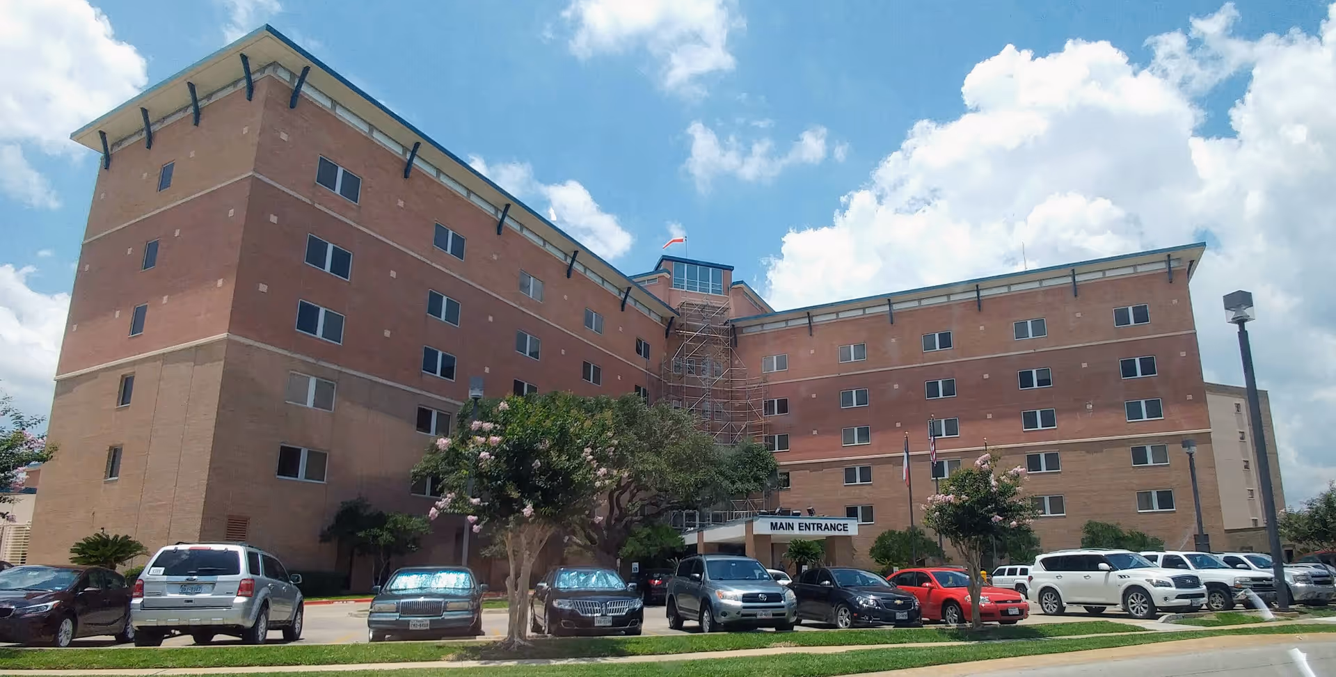 Brick medical center building with a main entrance, flagpoles, and a row of parked cars.
