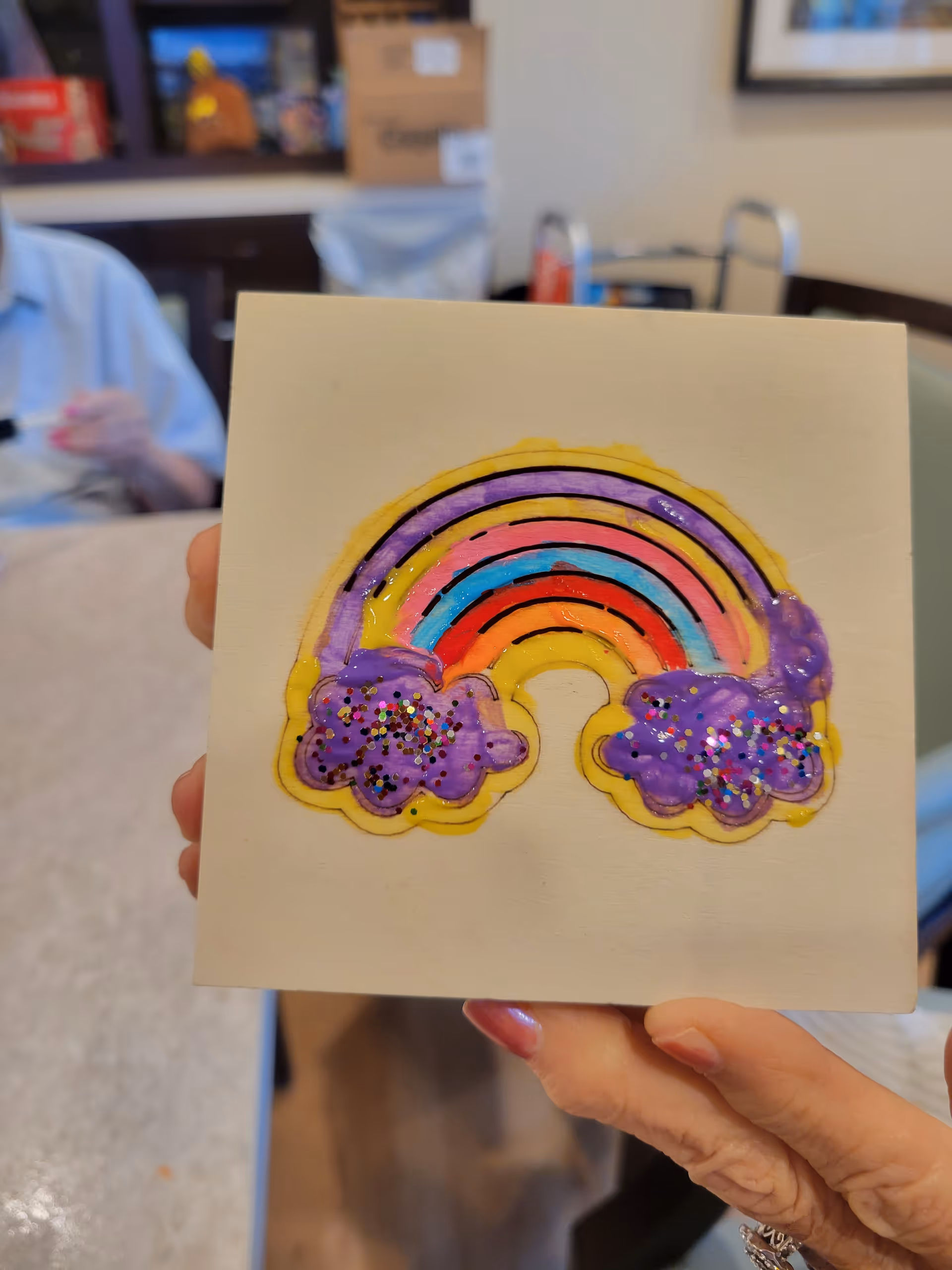 Hand holds a colorful rainbow artwork with purple clouds in a room.