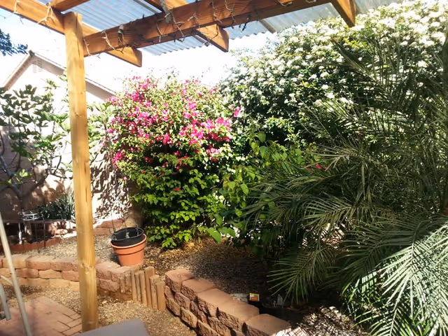Outdoor garden at a senior living facility featuring a wooden pergola, flowering shrubs, and potted plants.