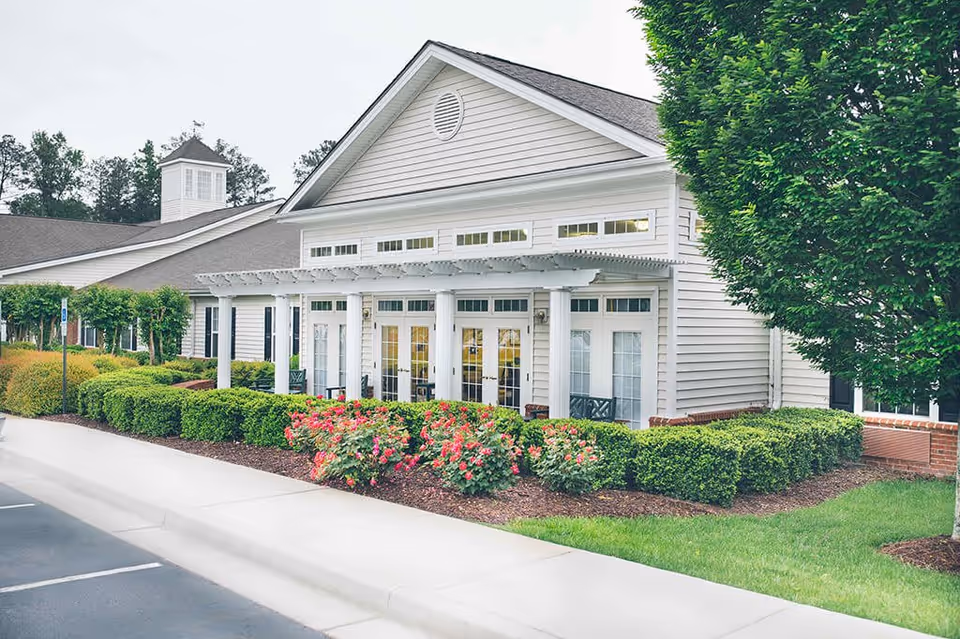 Exterior view of a white senior living facility building with a covered porch supported by columns, surrounded by neatly trimmed bushes and blooming pink flowers, with a parking lot in the foreground and trees in the background.