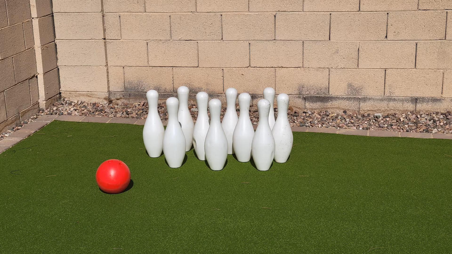 Outdoor artificial turf with white bowling pins lined up and a red ball nearby.