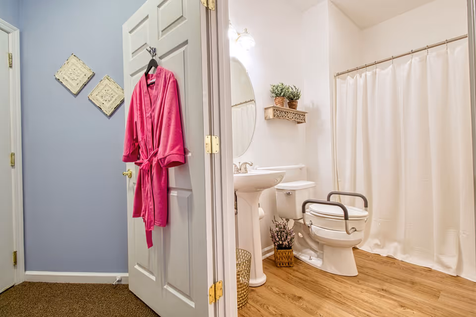Open bathroom with pedestal sink, toilet with grab bars, shower curtain, and pink robe.