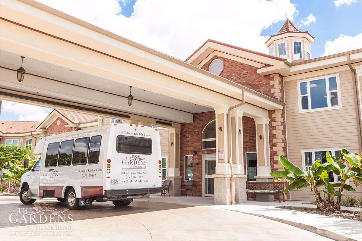 Entrance of The Gardens at Brook Ridge Assisted Living & Memory Care facility showing a covered drop-off area with a white shuttle van parked under the canopy. The building has beige siding with brick accents and large windows, and there are plants and a bench near the entrance.