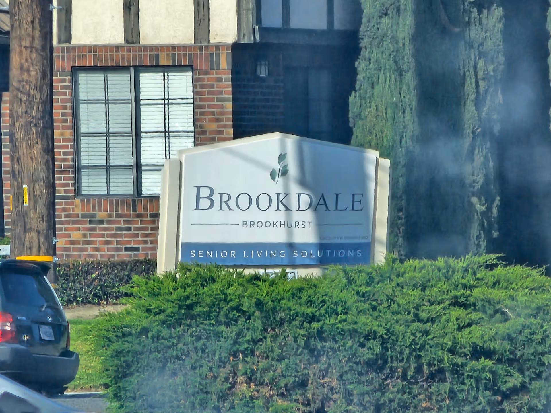 Brookdale Brookhurst sign on a brick building, surrounded by green bushes and a parked car.