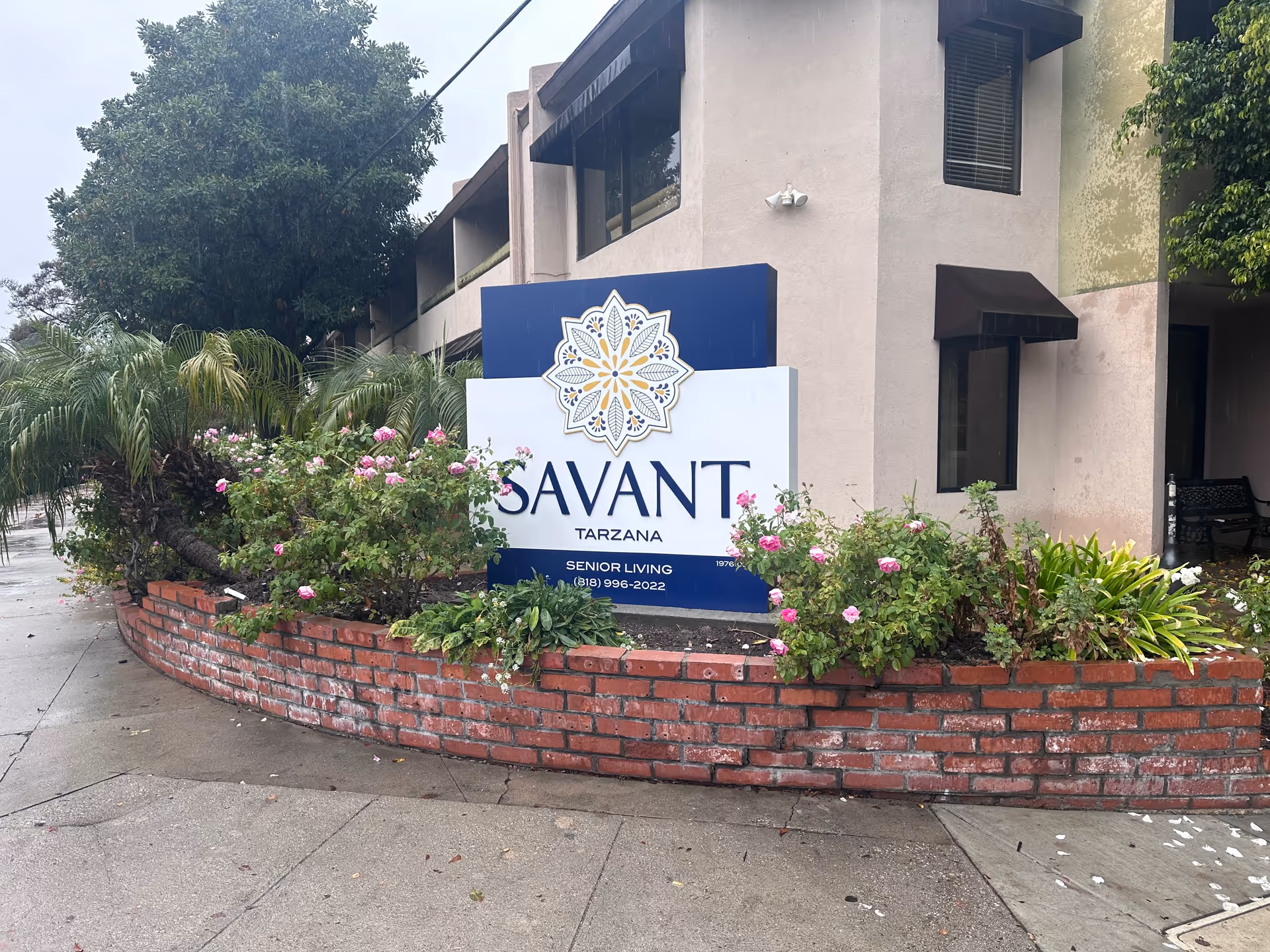 Exterior view of the Savant of Tarzana senior living facility sign surrounded by a brick planter with green plants and pink flowers, next to the building entrance.