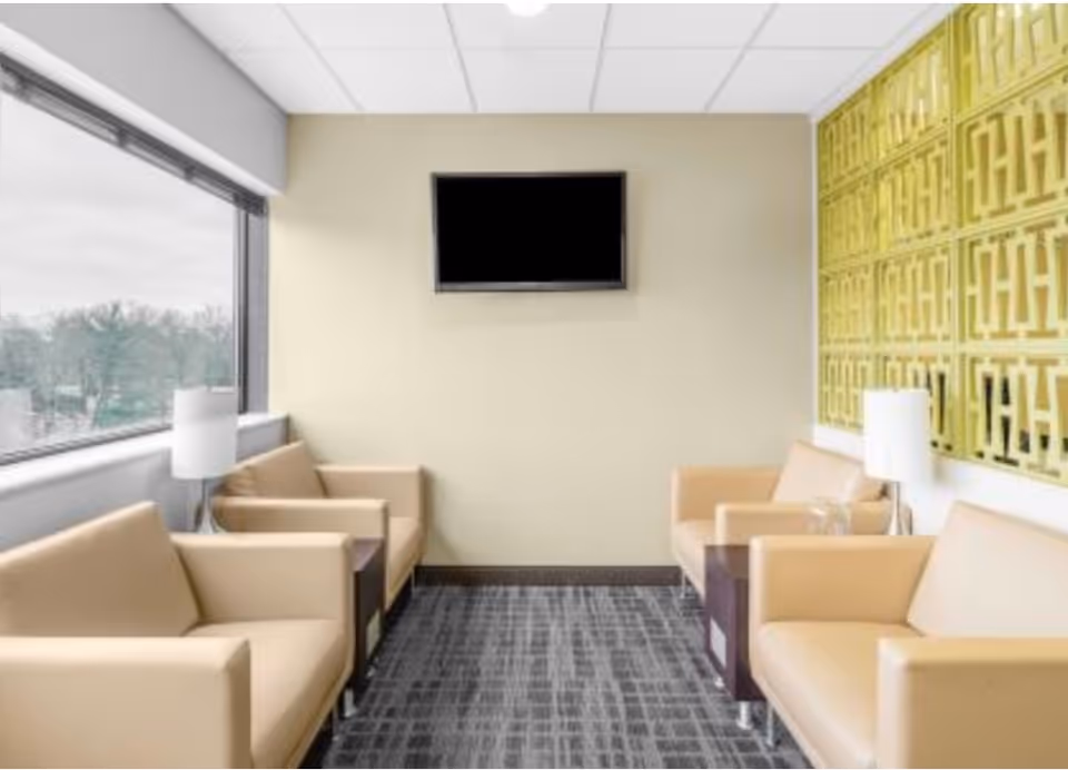 A modern senior living lounge with beige chairs, a TV, and a large window.