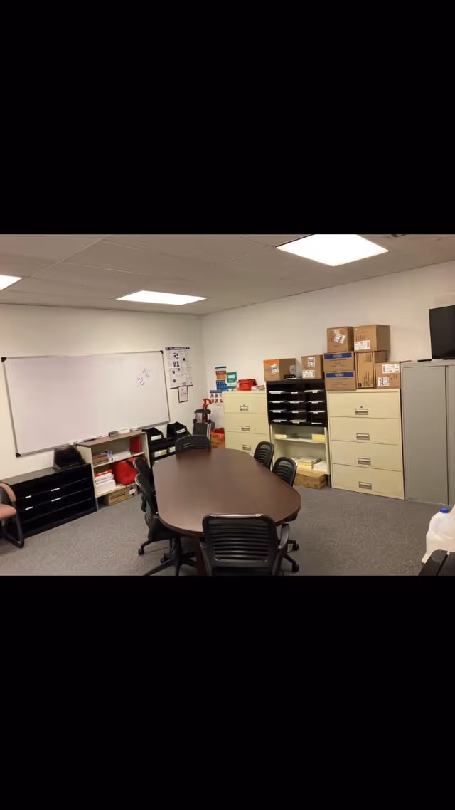 Oval conference table with chairs, whiteboard, and filing cabinets in a small meeting room.