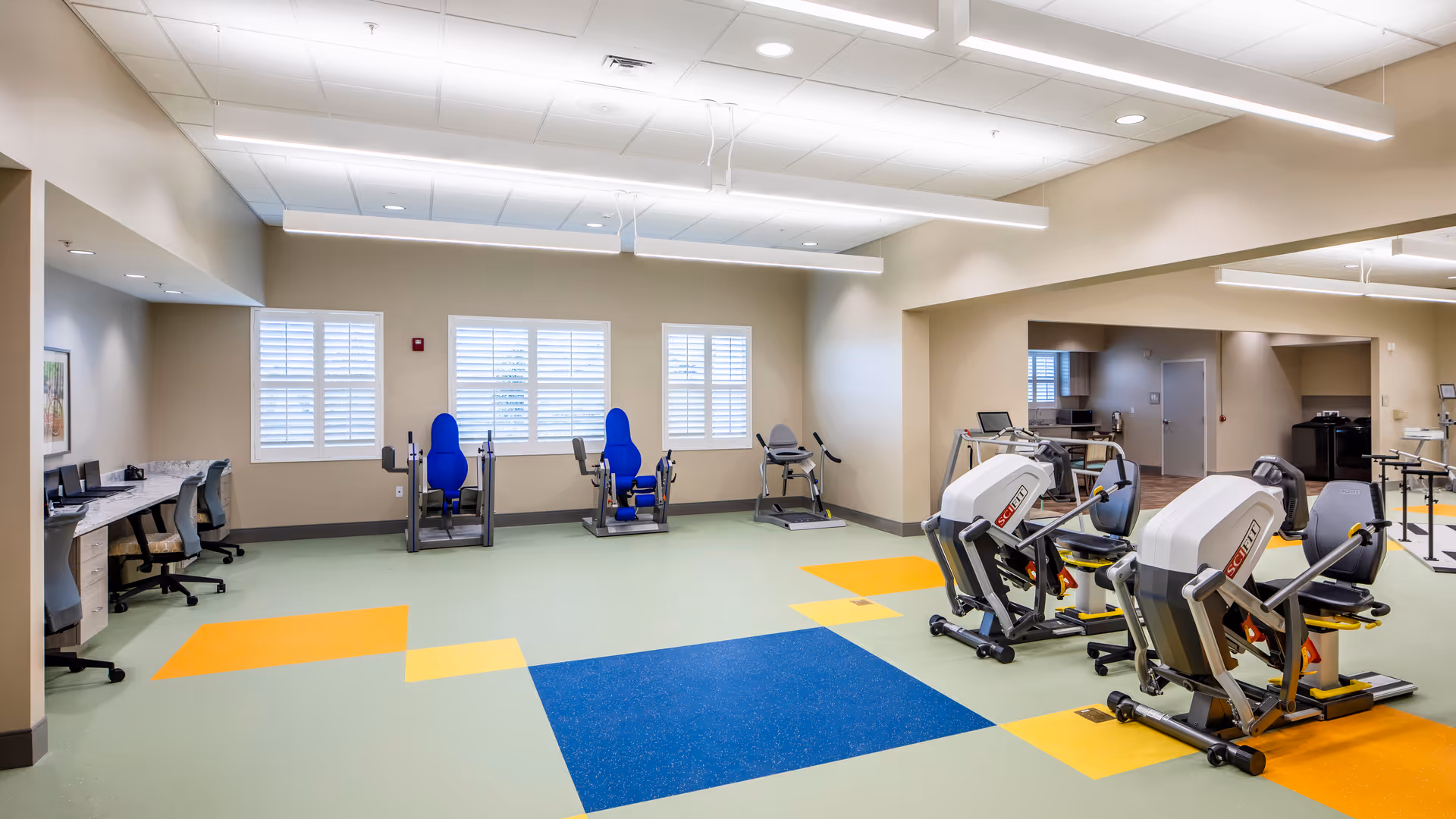 Bright rehabilitation gym with exercise machines, desks along the wall, and colorful tiled floor.