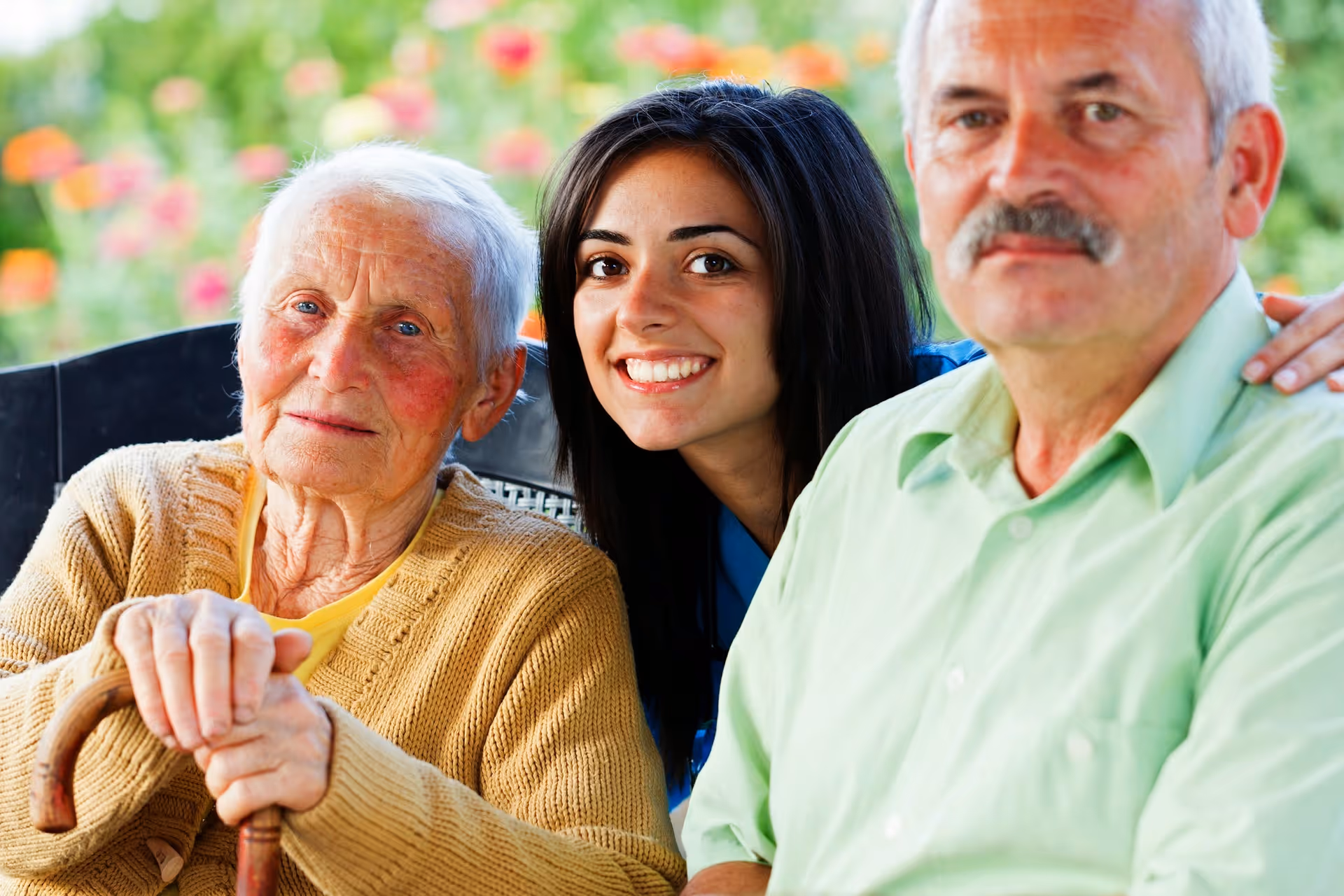 Three people outdoors, smiling together with an elderly woman holding a cane.