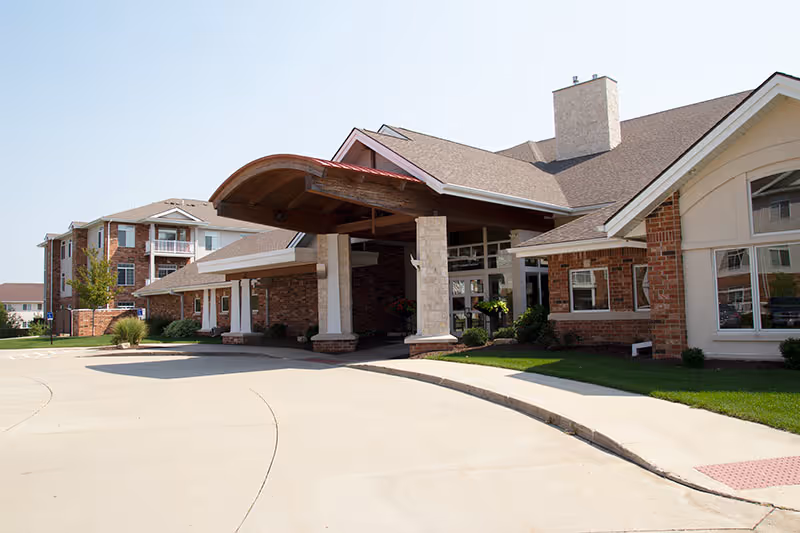 Exterior view of Woodlands Creek facility showing a large covered entrance with stone pillars and a curved wooden roof. The building is made of brick and has multiple windows. There is a paved driveway and some greenery around the entrance.